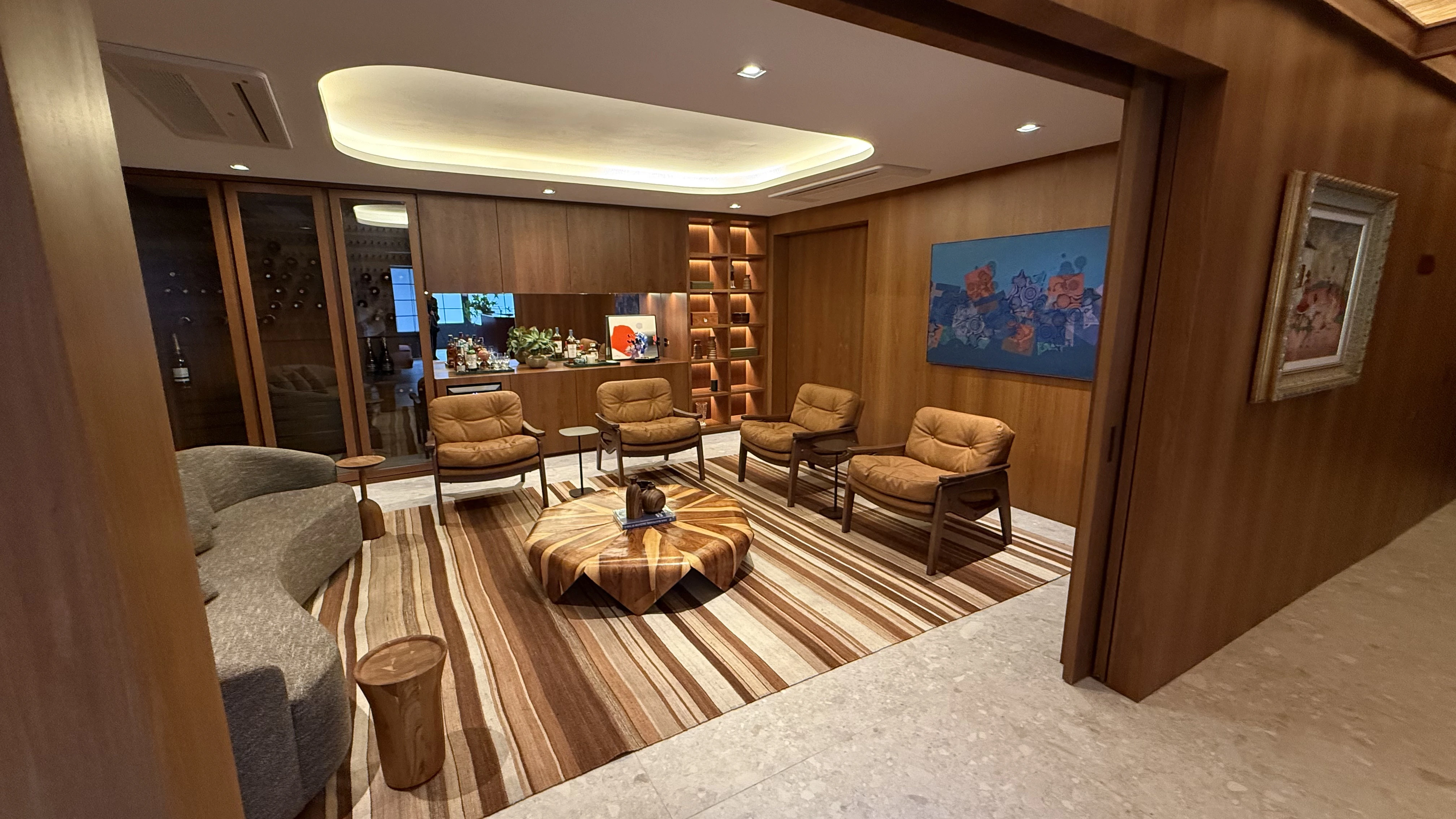 Lounge com bar revestido em marcenaria de madeira, poltronas de couro caramelo e iluminação indireta, projeto de Denise Kaupert